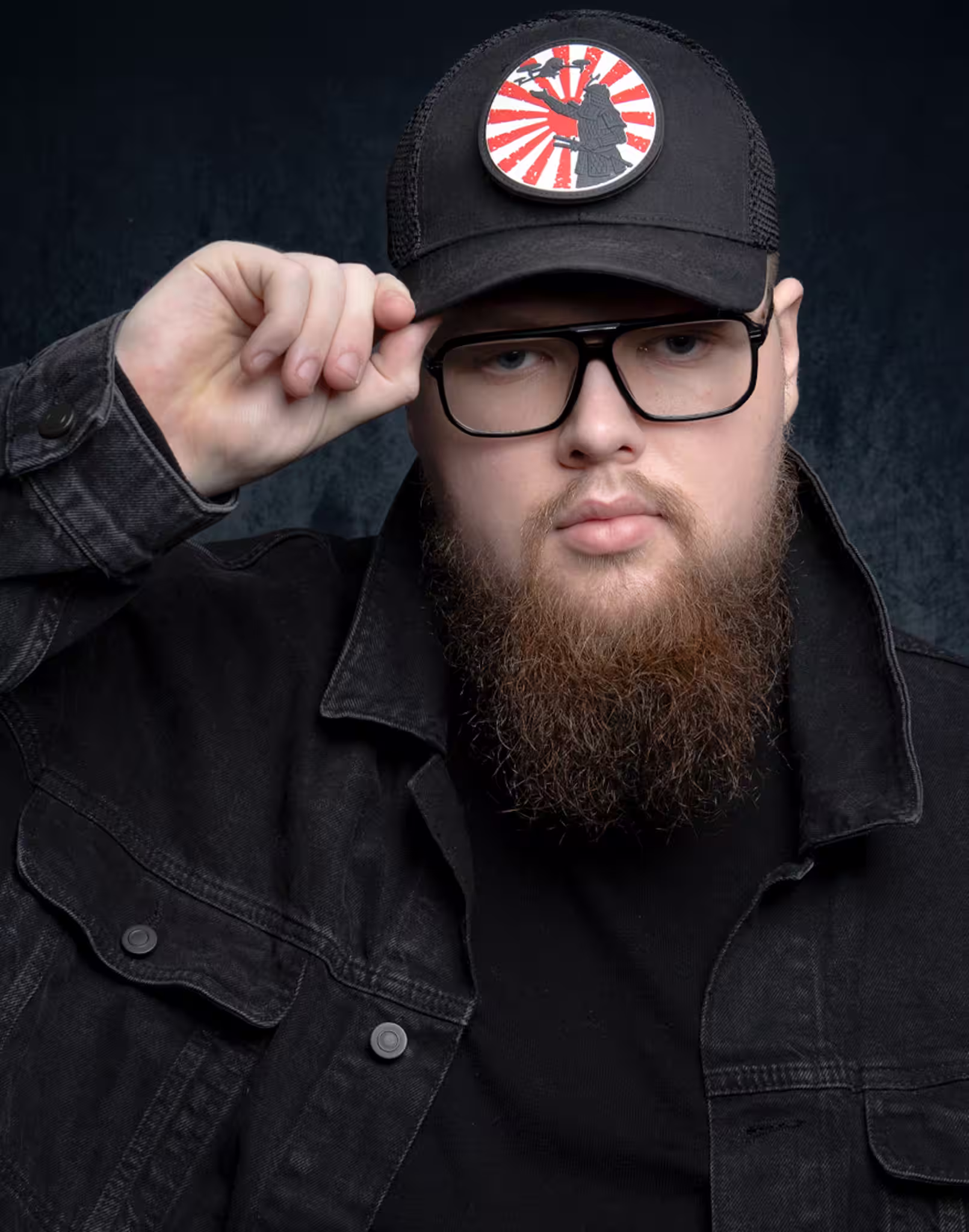 Fotograf headshot Leszno – autoportret Jakuba Lipnickiego, mężczyzny z brodą i okularami, w czapce z naszywką, trzymającego daszek, w ciemnej kurtce, na teksturowanym tle.